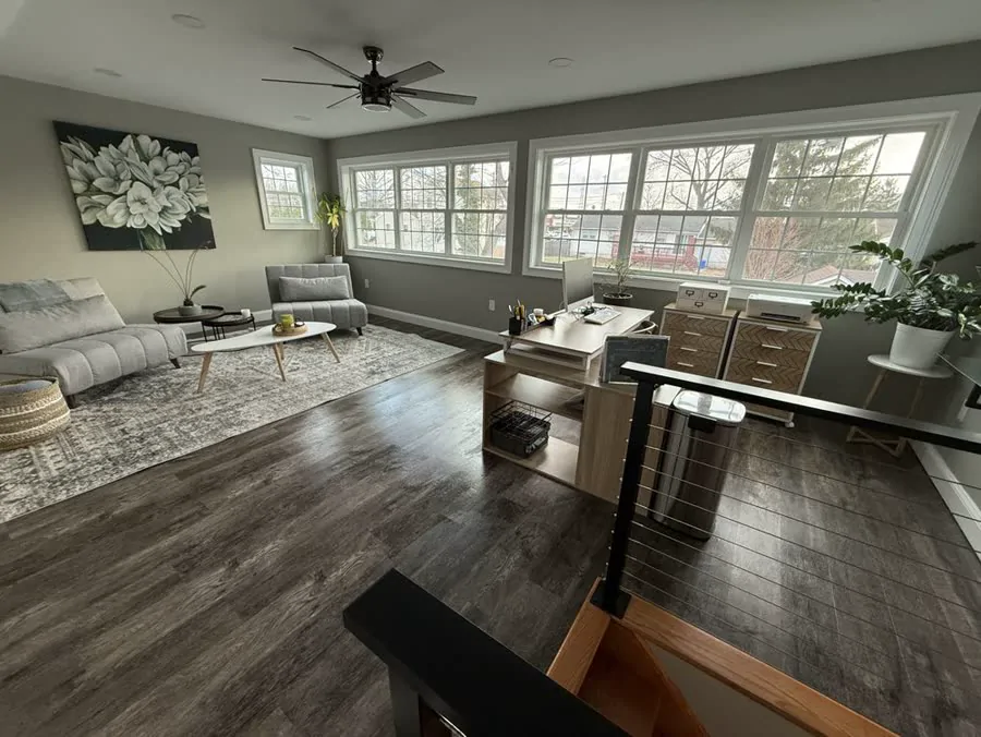 A wide view of a living room with a grey sofa, a ceiling fan, and big windows